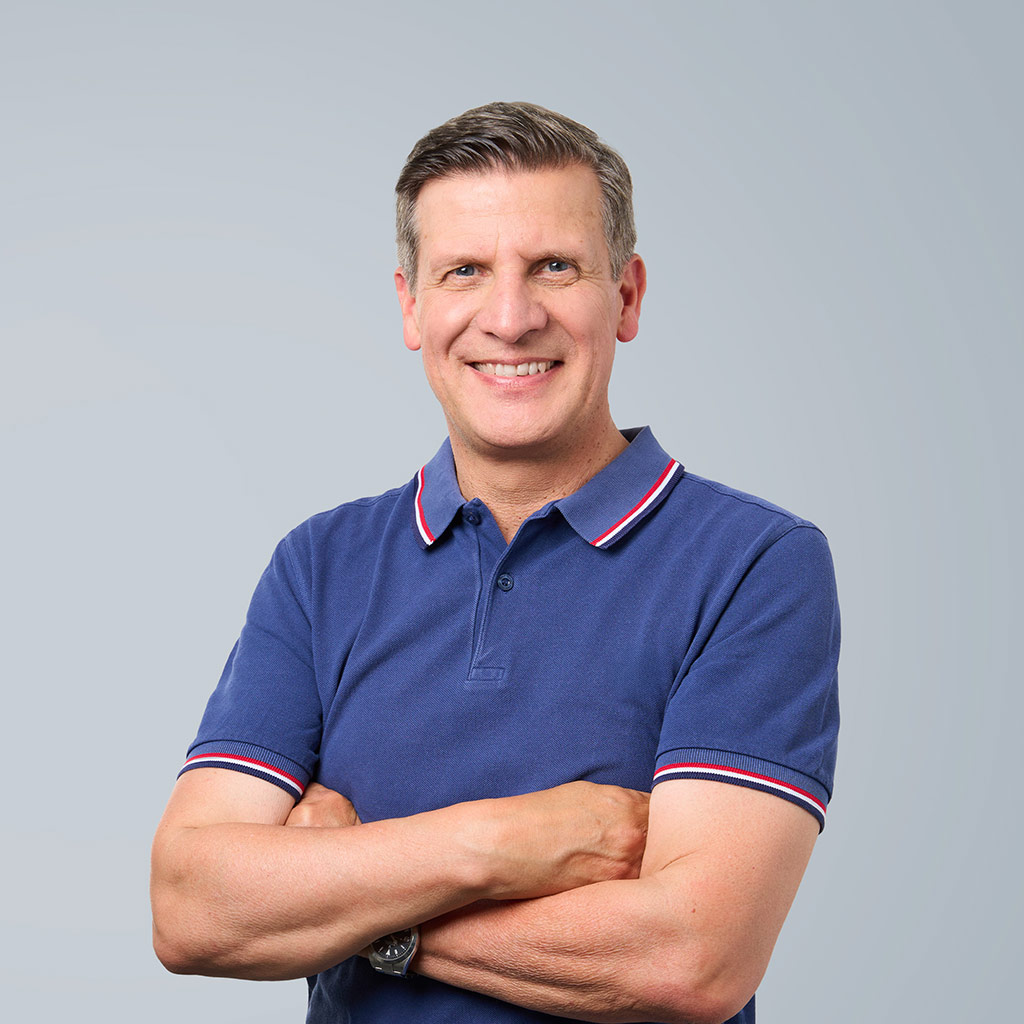 Lächelnder Mann mittleren Alters im blauen Poloshirt mit verschränkten Armen vor grauem Hintergrund.