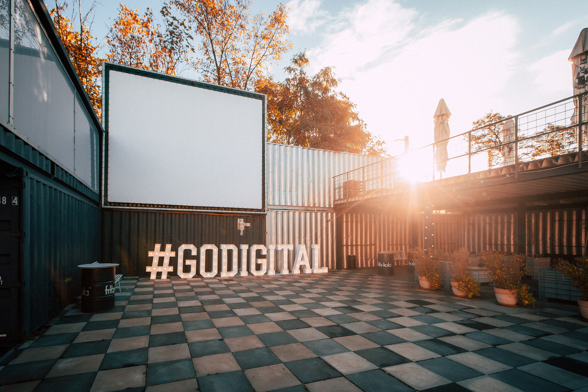 Eine Außenansicht bei Sonnenuntergang mit einem großen #GODIGITAL Schriftzug in leuchtenden Buchstaben vor gestapelten Schiffscontainern und einem karierten Boden.