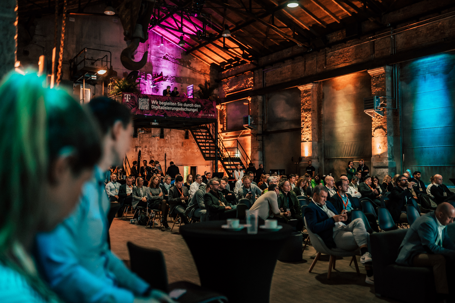 Menschen sitzen in einer großen, industriell aussehenden Halle bei einer Konferenz zum Thema Digitalisierung. Ein Banner im Hintergrund wirbt mit "Wir begleiten Sie durch den Digitalisierungsdschungel".