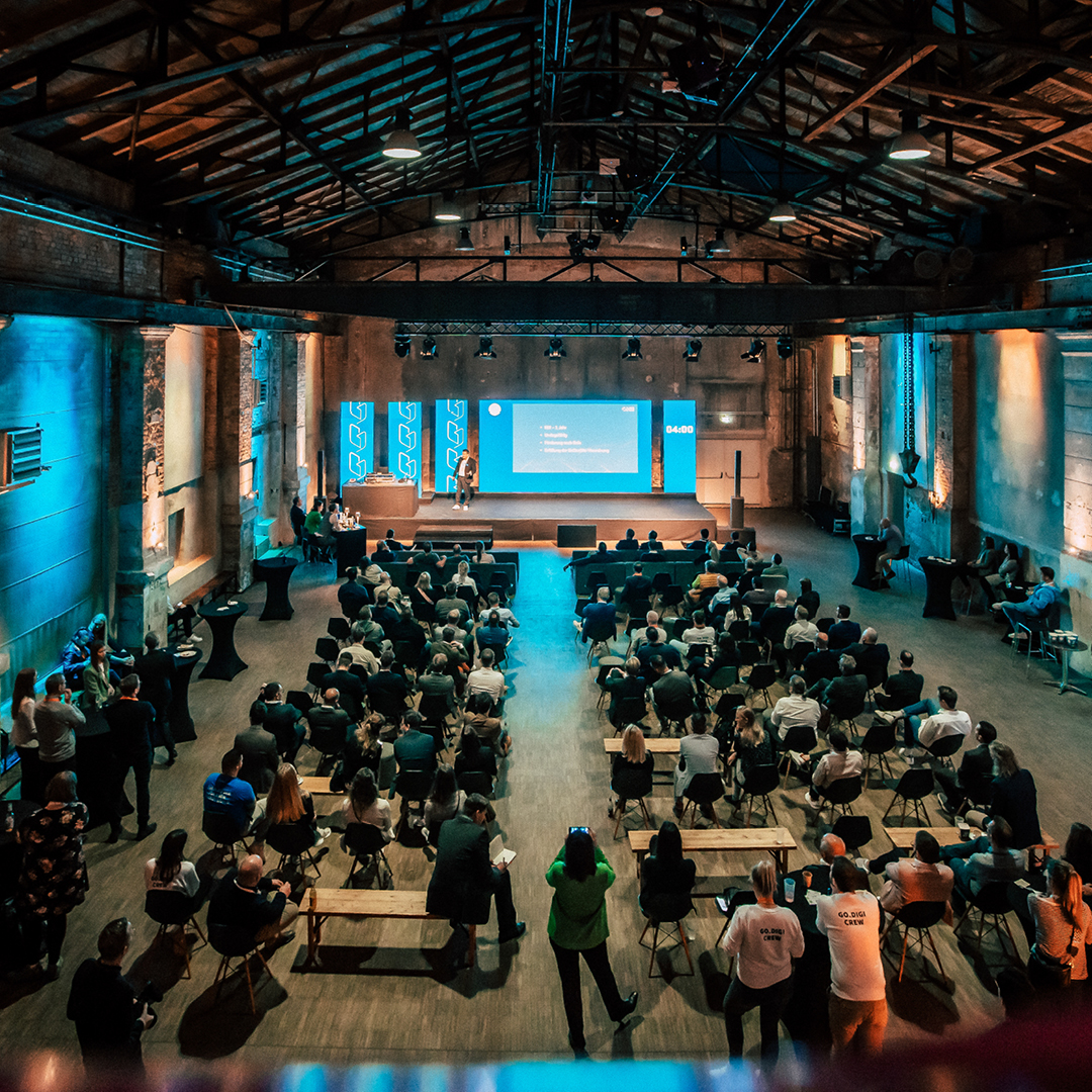 Konferenzsaal mit Publikum, das einer Präsentation über digitale Technologien in einem industriell gestalteten Raum folgt.