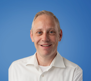 Porträt eines lächelnden mittelalterlichen Mannes mit kurzem blonden Haar, gekleidet in ein weißes Hemd vor einem blauen Hintergrund.