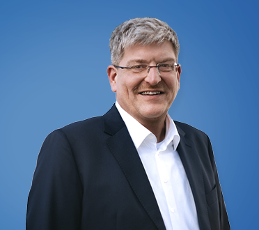 Porträt eines lächelnden Geschäftsmannes mit grauen Haaren, Brille und dunklem Anzug vor einem blauen Hintergrund.