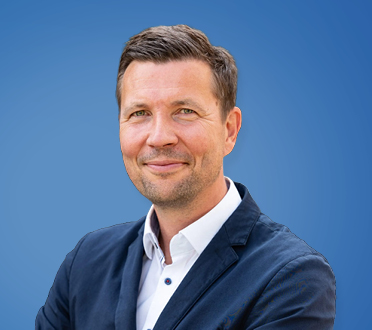 Porträt eines lächelnden Geschäftsmannes in Anzug und Hemd vor einem blauen Hintergrund.