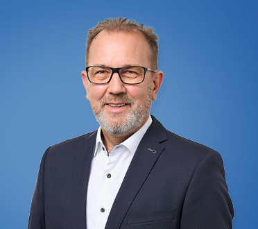 Porträt eines lächelnden Geschäftsmannes mit Brille und grauem Haar, gekleidet in ein dunkelblaues Sakko und weißes Hemd vor einem blauen Hintergrund.