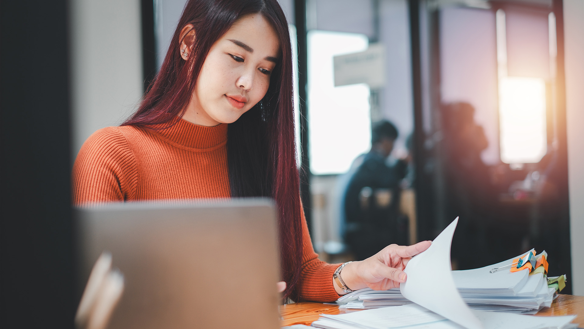 Eine Frau mit langen, roten Haaren arbeitet konzentriert an einem Tisch, umgeben von Dokumenten und einem Laptop.