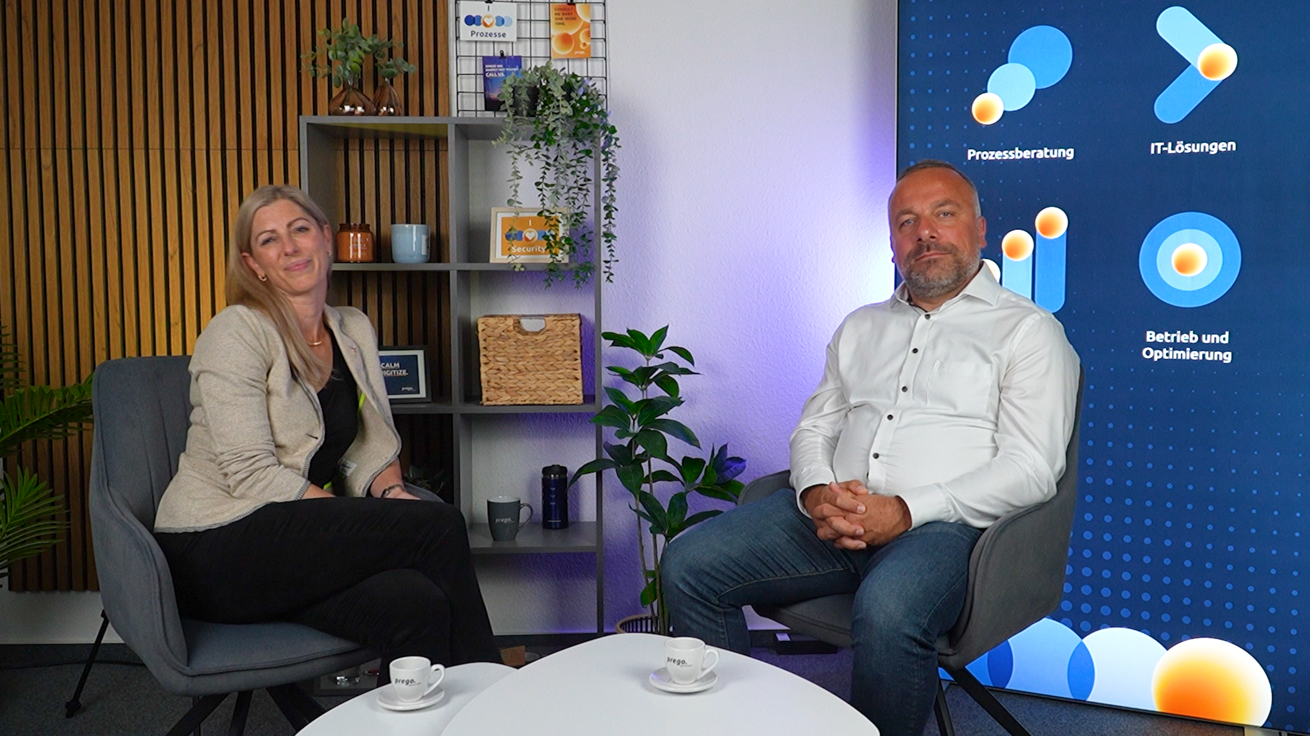 Webinar über IT-Lösungen für Versorger Zwei Personen sitzen in einem modernen Büro, umgeben von Pflanzen und Regalen. Im Hintergrund sind Grafiken zu IT-Lösungen und Prozessberatung sichtbar.